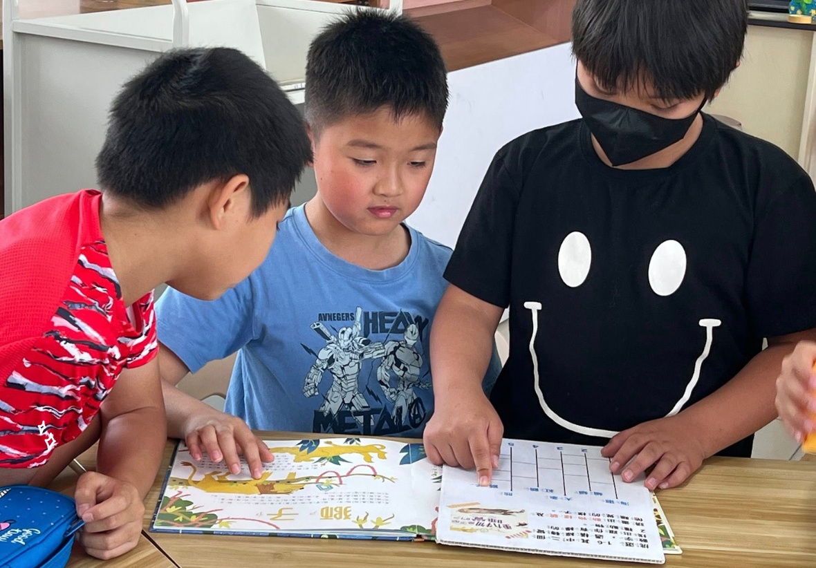 三名小學生圍坐在木製桌前，專注地查看打開的書籍和資料。左邊男童穿紅色條紋衣，中間男童穿藍色漫威T恤，右邊男童穿黑色笑臉圖案T恤並戴著黑色口罩。他們正在進行閱讀或學習活動，室內為教室環境。