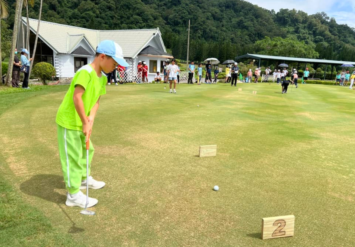 一名穿著螢光綠色衣服、戴著藍色帽子的年輕男孩在高爾夫球場上揮桿，準備打第二洞。背景是白色clubhouse建築、觀眾群眾和樹林，這是一場社區高爾夫球賽事。