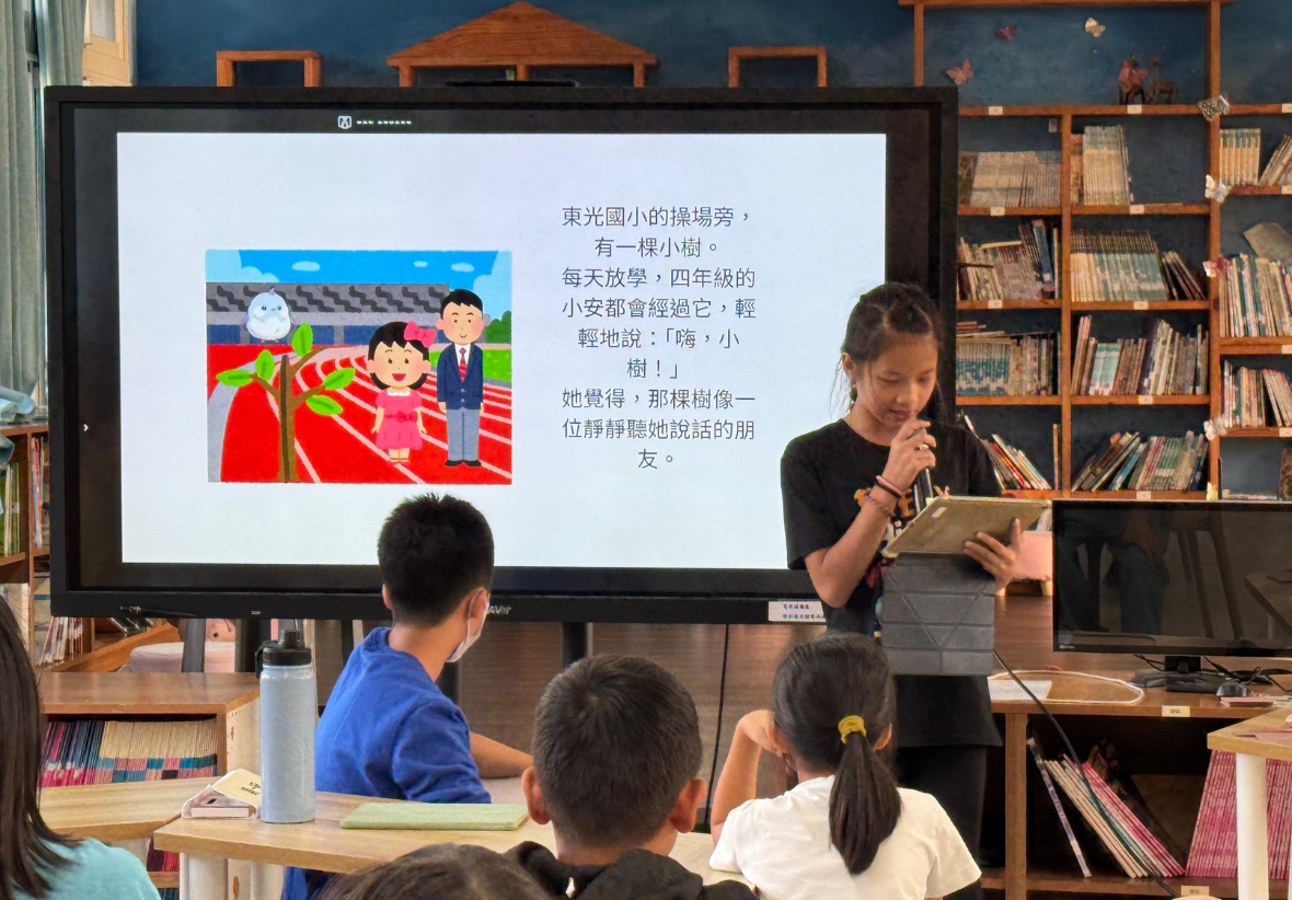 圖書館內，教師手持書本站在大螢幕前為四名學童進行中文故事讀書會。螢幕上顯示插圖和繁體中文文字，講述關於東光國小操場旁小樹的故事。背景為木製書架，環境溫暖舒適。