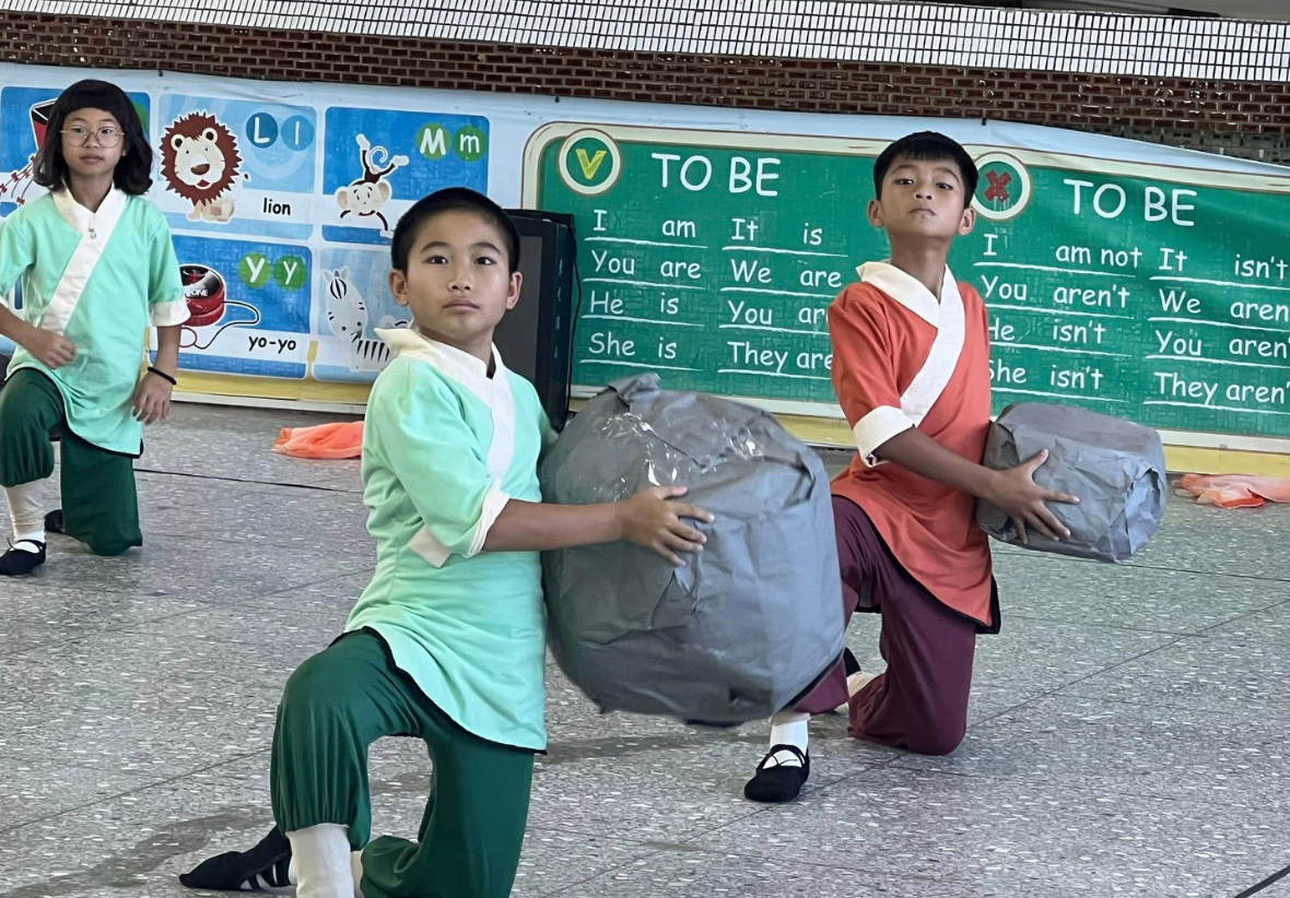 教室內，三位穿著傳統服飾的學童跪坐在地上，正在進行英語課程活動。他們各自抱著灰色布袋，專注地看向黑板。黑板上展示「TO BE」動詞的各種變化形式。左側牆上貼著字母學習卡。後方另有一位學童站立。