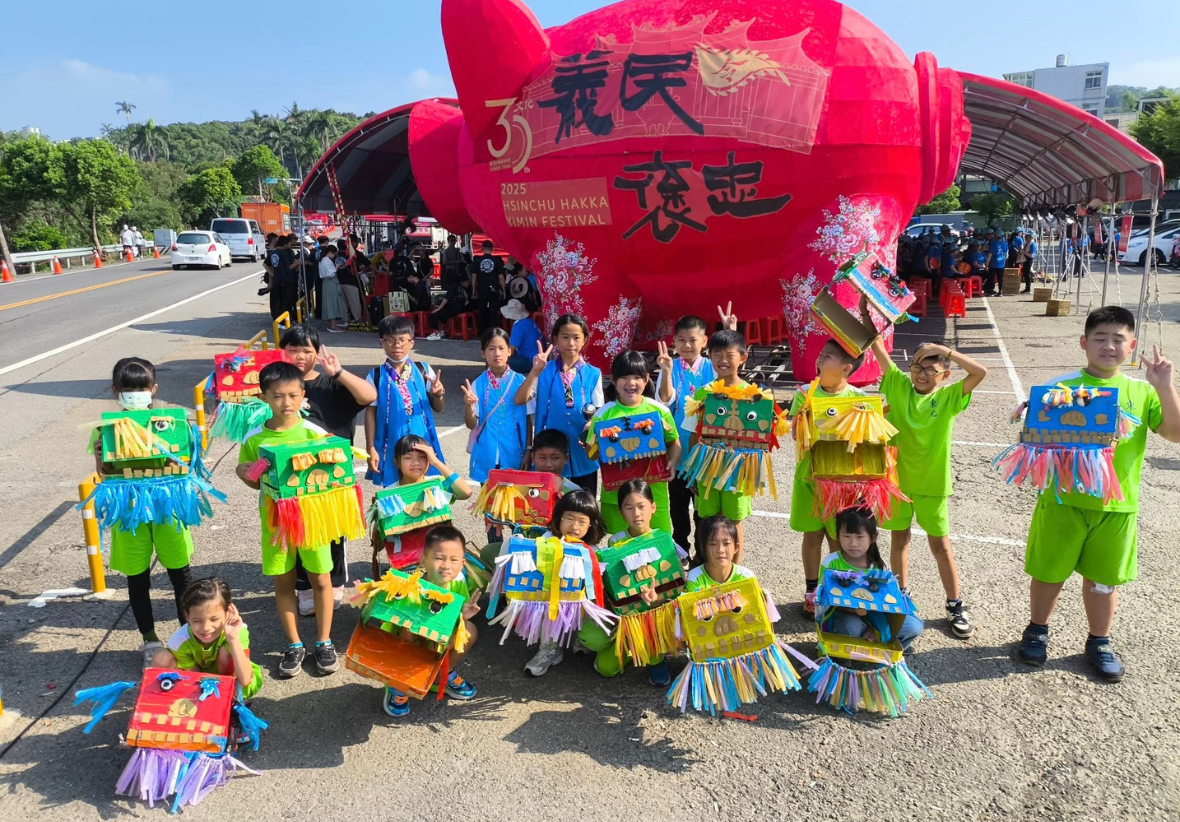 新竹客家旗袍節慶活動現場，一群身穿彩色傳統服飾的兒童在大型紅色燈籠布景前合照。背景有遊客、帳篷和停泊的車輛，展示客家文化節慶的熱鬧氛圍。