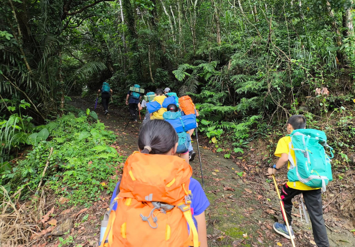 一群登山者穿著彩色背包和登山裝備，在茂密的熱帶森林中沿著泥土小徑行進。前景的登山者穿著橙色背包，隊伍依序跟進，四周被高大樹木和茂盛植被包圍，呈現叢林登山探險的情景。