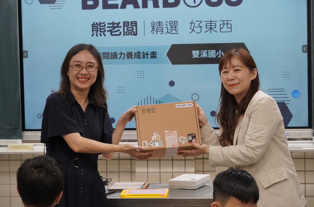 數位大螢幕前，左側穿黑連身裙的女性校方代表與右側穿條紋西裝的企業代表（來自BEARBOSS 熊老闆），共同雙手交接一個貼有貓咪等可愛貼紙的牛皮紙箱禮盒。兩人對鏡頭展現燦爛喜悅的笑容，氣氛溫馨。