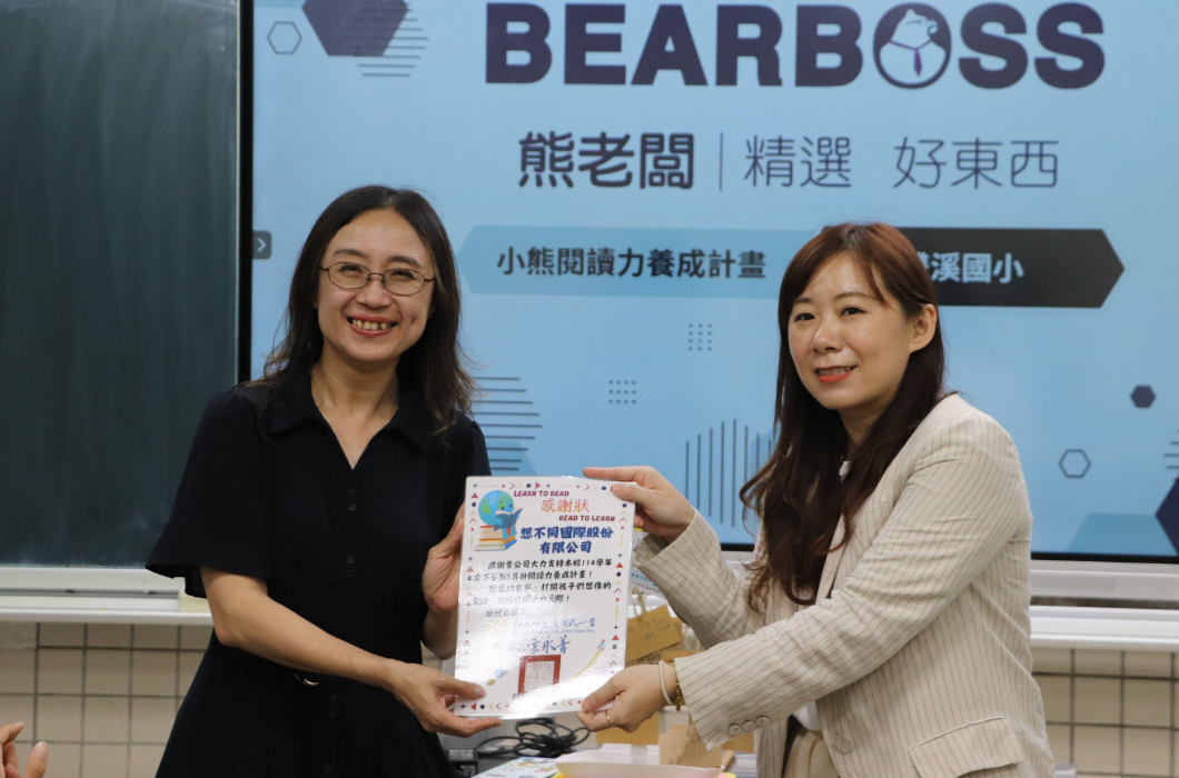 承接上一張場景，兩位女性代表共同舉起一張裝飾精美的官方「感謝狀」向鏡頭展示。感謝狀上寫著「感謝想不同國際股份有限公司（熊老闆）大力支持本校114學年度小熊閱讀力養成計畫」，雙方臉上充滿欣慰的笑容。