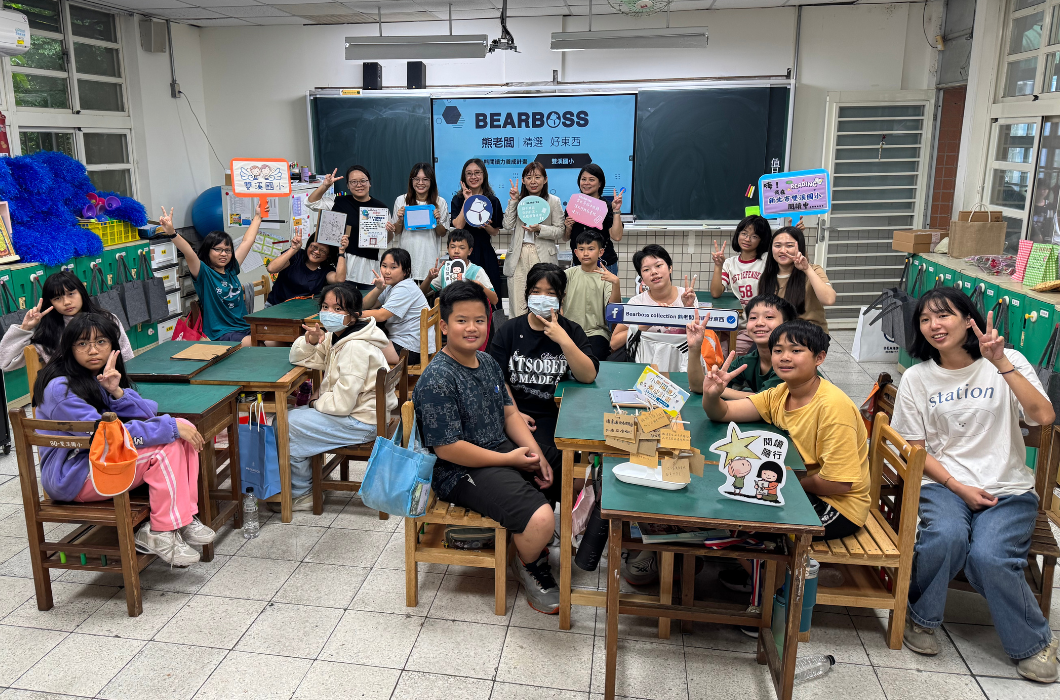 明亮的教室裡，全體國小學童與講師、企業夥伴及師長們開心圍聚在一起大合影。前排與兩旁的孩子們紛紛比出勝利手勢（V），後方的師長們則舉著繽紛的宣傳對話框手牌。每個人臉上都洋溢著滿滿的笑容與幸福感。