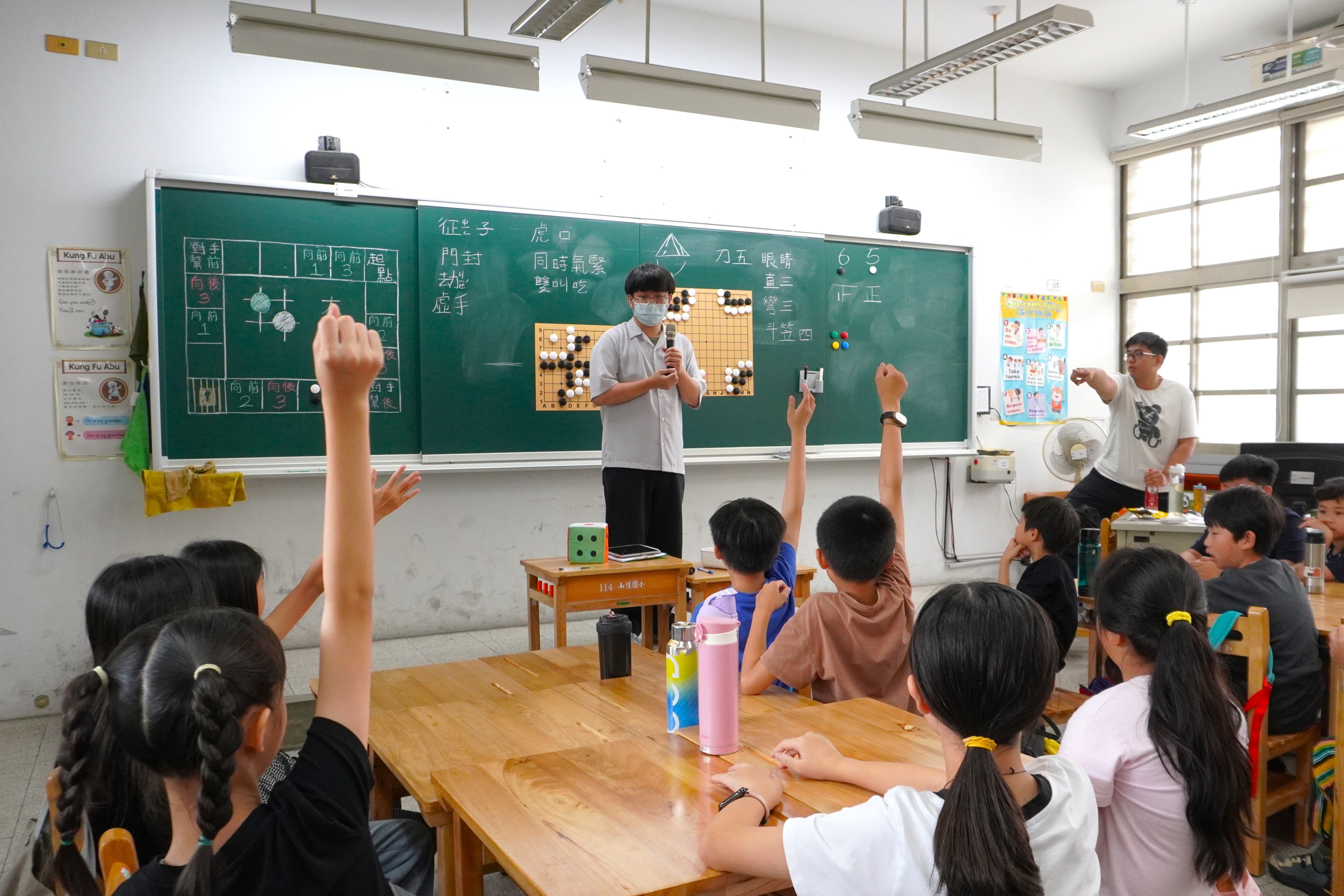 圍棋課堂中，戴口罩的青年站在黑板前拿著麥克風講解，多名學生坐在木桌旁舉手回應，另一名青年在旁協助引導。