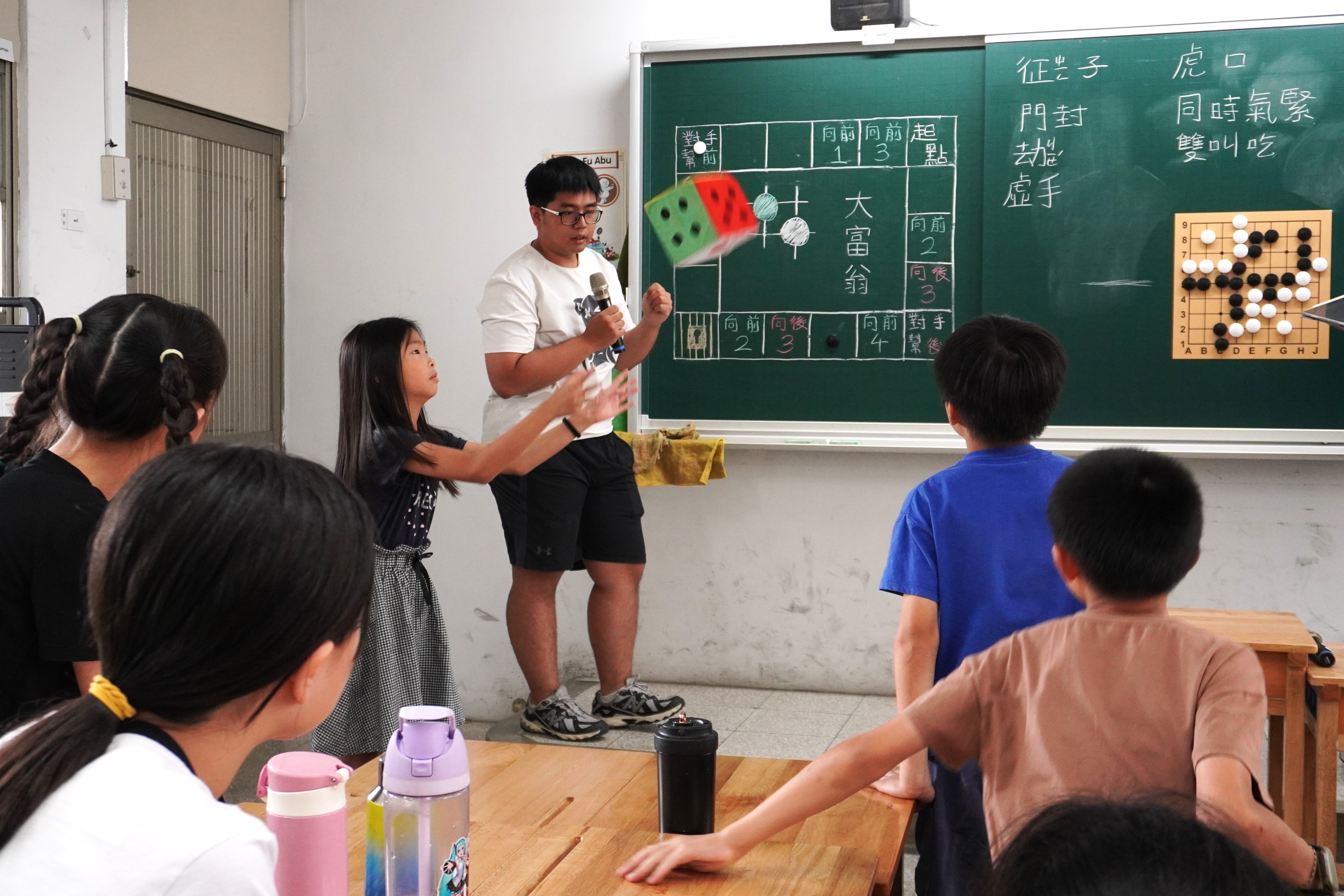 教室前方，一名青年手持麥克風帶領遊戲，女孩將大型彩色骰子拋向空中，其他學生坐在座位上注視，黑板旁掛著圍棋盤。
