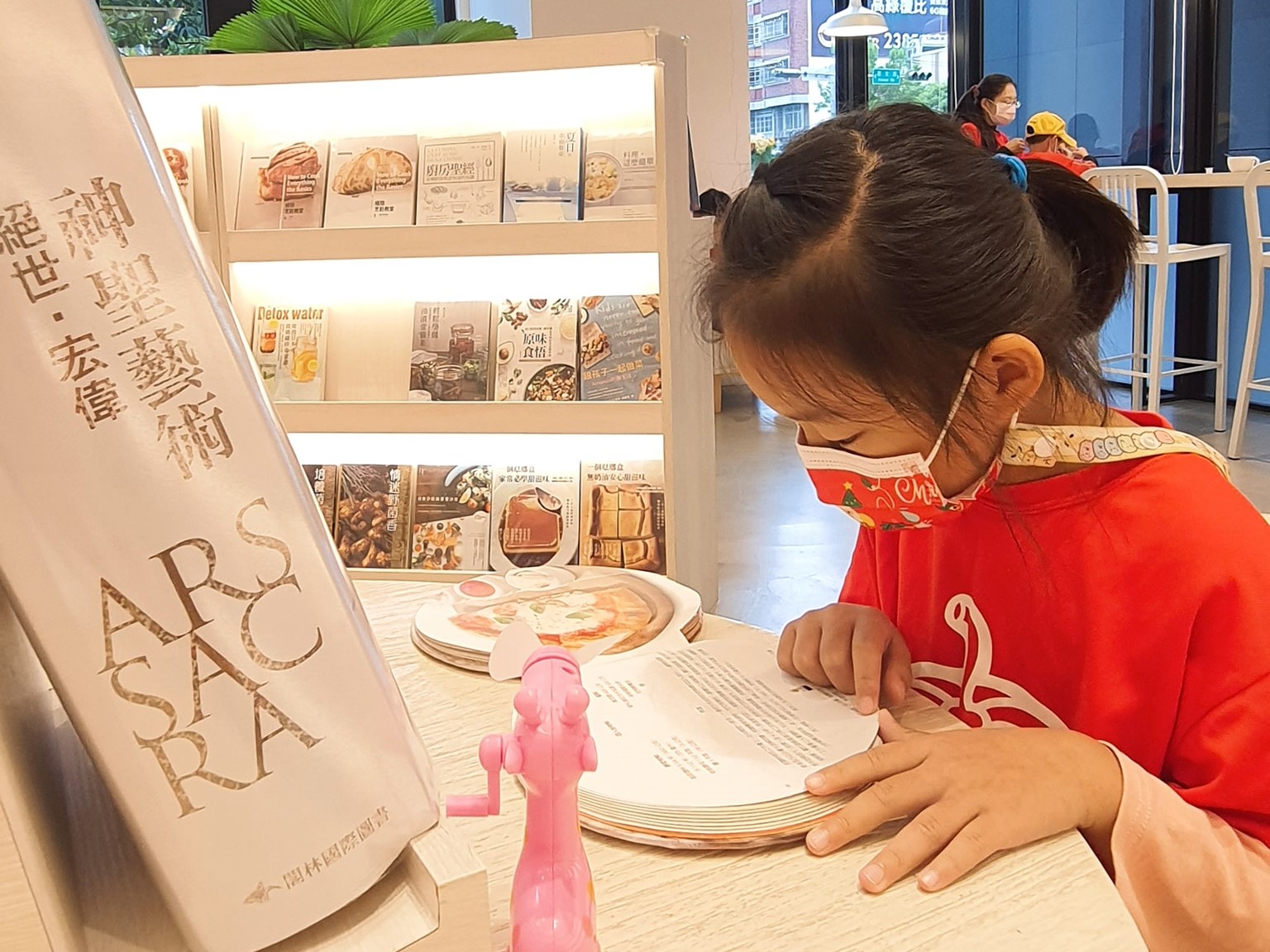 小女孩穿著紅色衣服，在室內展示區查看美食相關的互動展覽。桌上放著印有食物圖案的盤子和粉紅色玩具。背後是三層展示架，陳列各式餐飲宣傳冊。左側立牌展示「SACS PARK」字樣及中文文字。