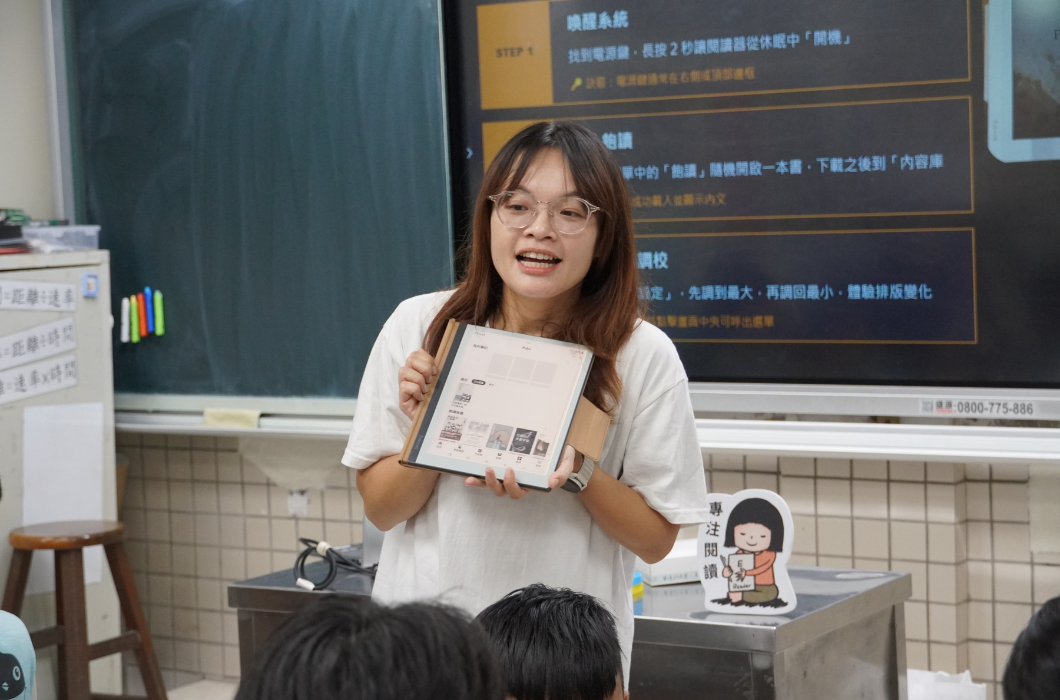 課堂前方，一位戴眼鏡、穿白上衣的長髮女性講師，正雙手拿著一台開啟的電子書閱讀器展示給台下的學生看。她身後的數位大螢幕上顯示著「喚醒系統」、「飽讀」與「調校」等使用步驟說明，一旁桌上還放著「專注閱讀」的小立牌。