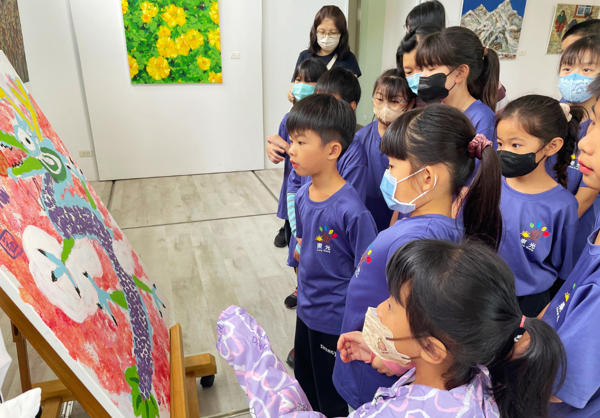 一群穿著紫色制服的學童在美術館欣賞展覽作品，大部分學生戴著口罩。他們聚集在一幅大型彩色畫作前，該作品以紅色為主，有白色人物和綠色裝飾元素。背景牆面上掛著黃色花卉和山景畫作。