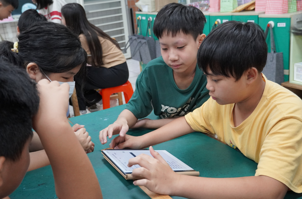 三位男同學圍繞著綠色課桌。坐在右側穿黃色上衣的男孩一邊看著電子書閱讀器上的選單，一邊伸出手指在螢幕上操作。中間穿綠色上衣的男孩與左側的同學則湊近觀看，神情認真，正互相討論、研究如何操作系統。