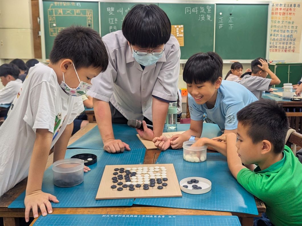 圍棋桌旁，戴口罩的青年拿著麥克風俯身觀察棋局，三名男孩圍在棋盤四周思考，其中一人面露笑容，桌上擺滿黑白棋子。