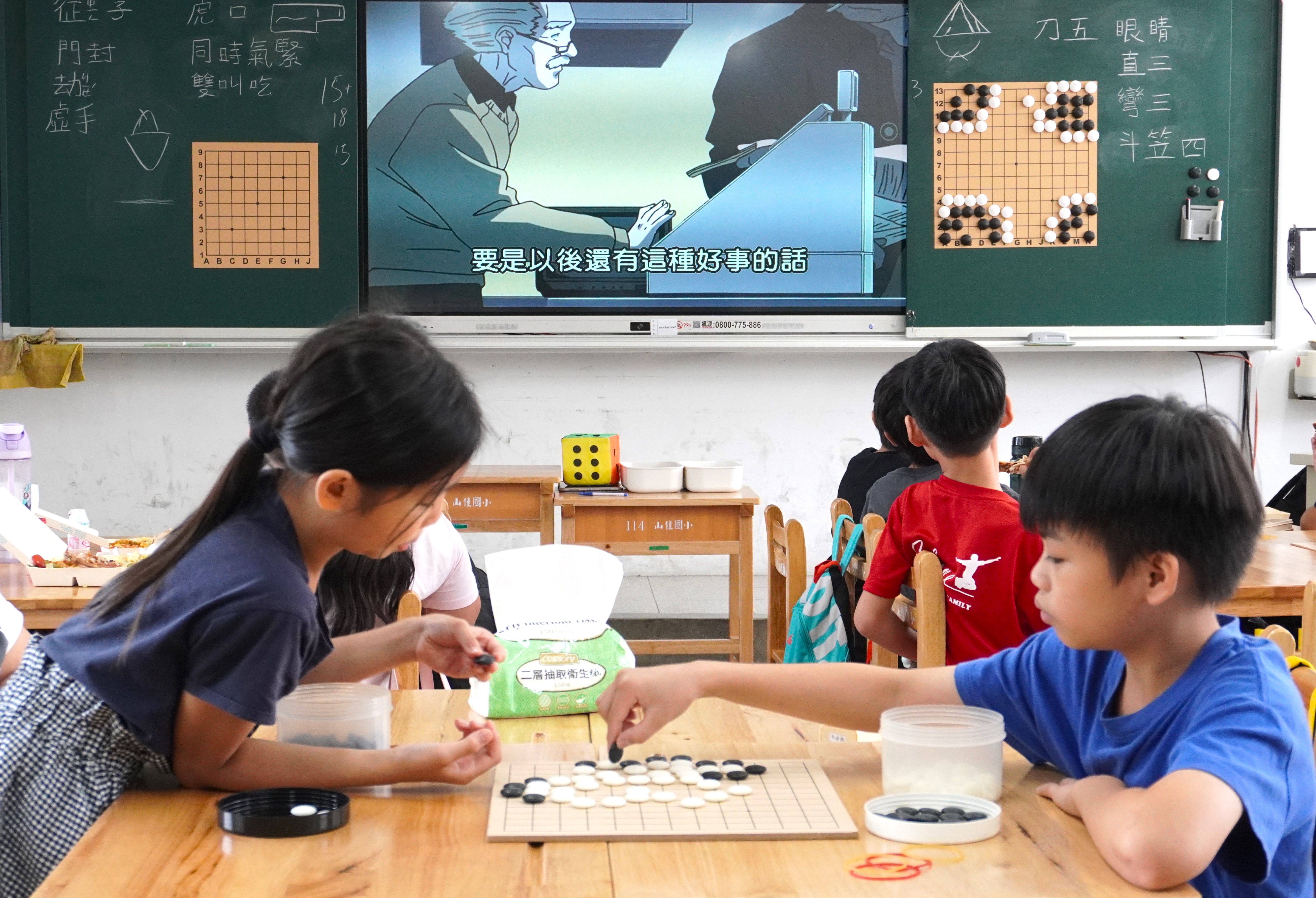 教室內，兩名學生在前方木桌對弈，手持黑白棋子思考落子；後方學生面向電子白板觀看動畫《棋靈王》，黑板兩側掛有圍棋盤。