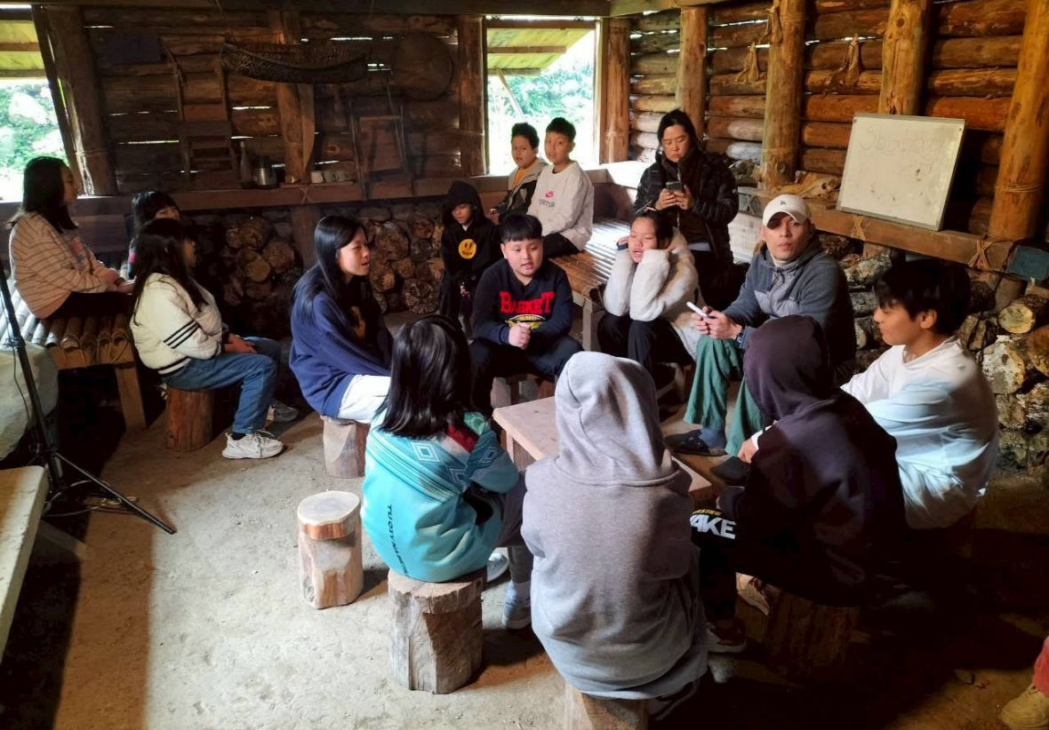 在充滿原住民傳統特色的木造家屋內，牆邊堆滿薪柴，學生與老師們圍坐在矮木凳與竹榻上進行小小的部落會議。大家面對面聚在一起認真討論與交流，氣氛專注而溫馨。