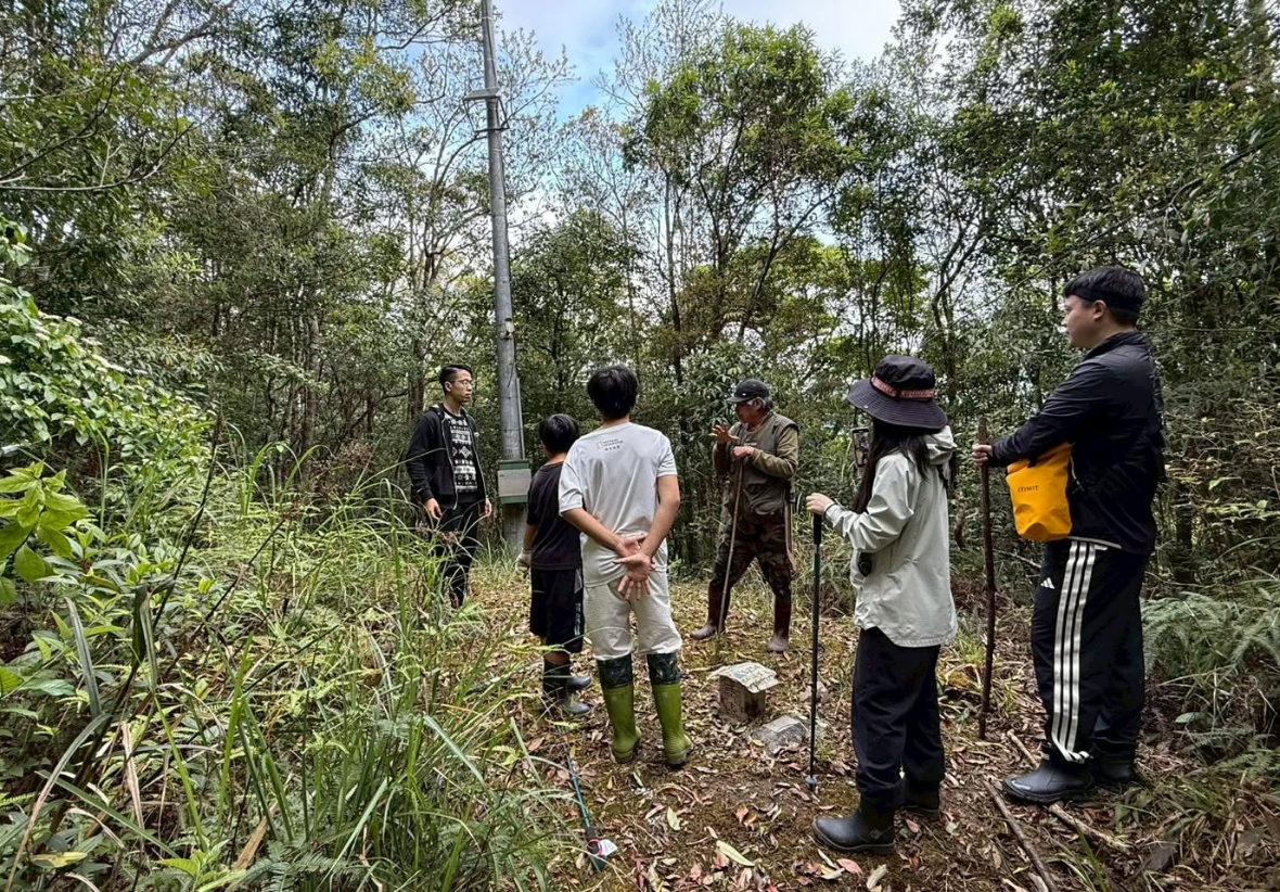 在茂密的山林步道中，部落長者手持竹杖，向圍繞在身邊的師生與青年進行山林植物與生態文化的導覽解說。參與者們身穿戶外服裝、腳踩雨鞋，認真聆聽，深刻體驗自然山林的智慧。