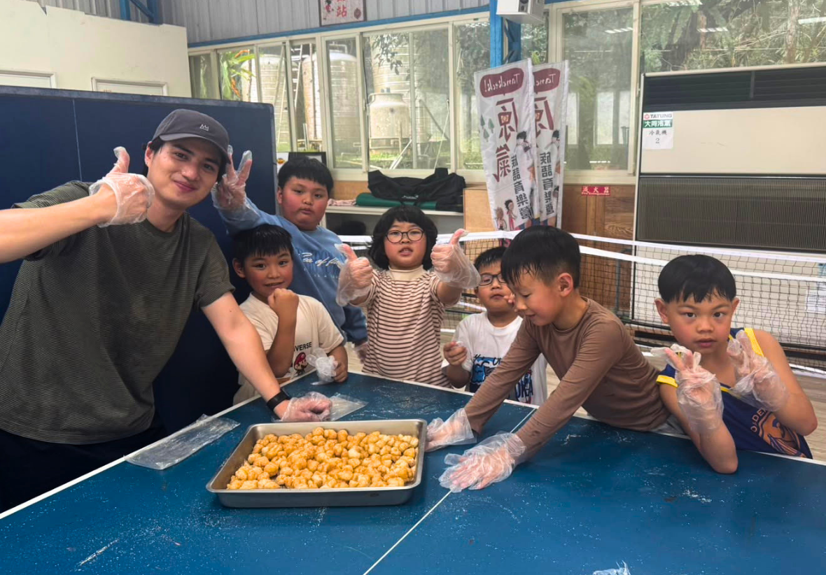 在桌球桌前，一位男老師與六位國小學童戴著透明手套，歡樂地圍在一大盤剛製作完成、裹滿花生粉的「小米麻糬」旁。大家開心對著鏡頭比出讚或勝利手勢，展現親手製作原鄉傳統美食的滿滿成就感與歡笑。