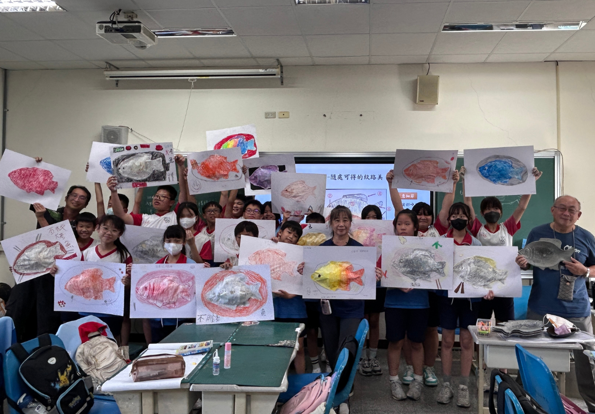 一個教室裡，大約三十位學生和教師舉著自己繪製的魚類插畫，展示各種顏色和形態的魚。黑板上寫著中文字，牆上貼著更多魚類繪畫作品。這是一場海洋生物主題的美術教學活動展示。