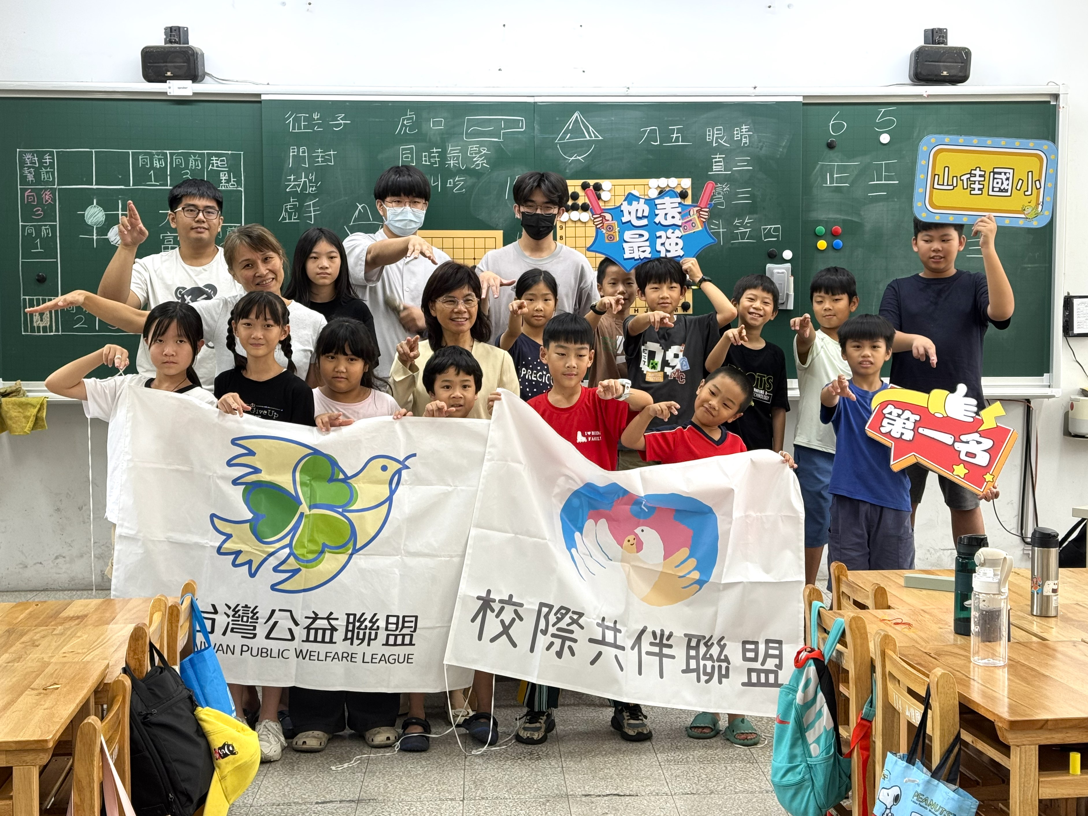 師生在教室黑板前合影，眾人比出手勢，前排拉起「台灣公益聯盟」與「校際共伴聯盟」布條，山佳國小學生手持標語牌。