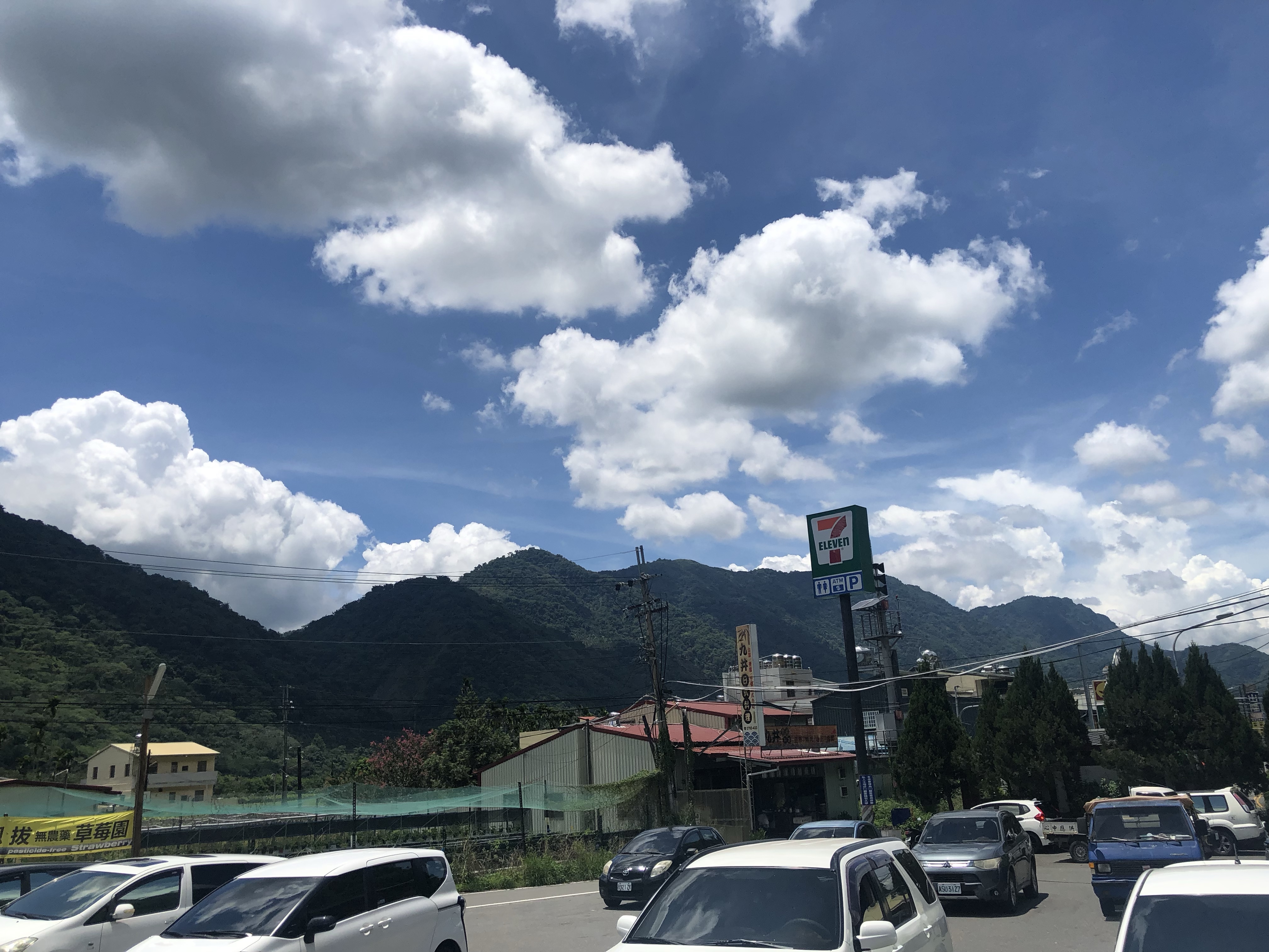 山區停車場景，7-Eleven便利店招牌立於停車場旁，多輛汽車停放其中。背景為青翠的群山環繞，藍天白雲，山腰上可見工業建築。電線和路標散布周圍，呈現典型的日本鄉村小鎮風景。
