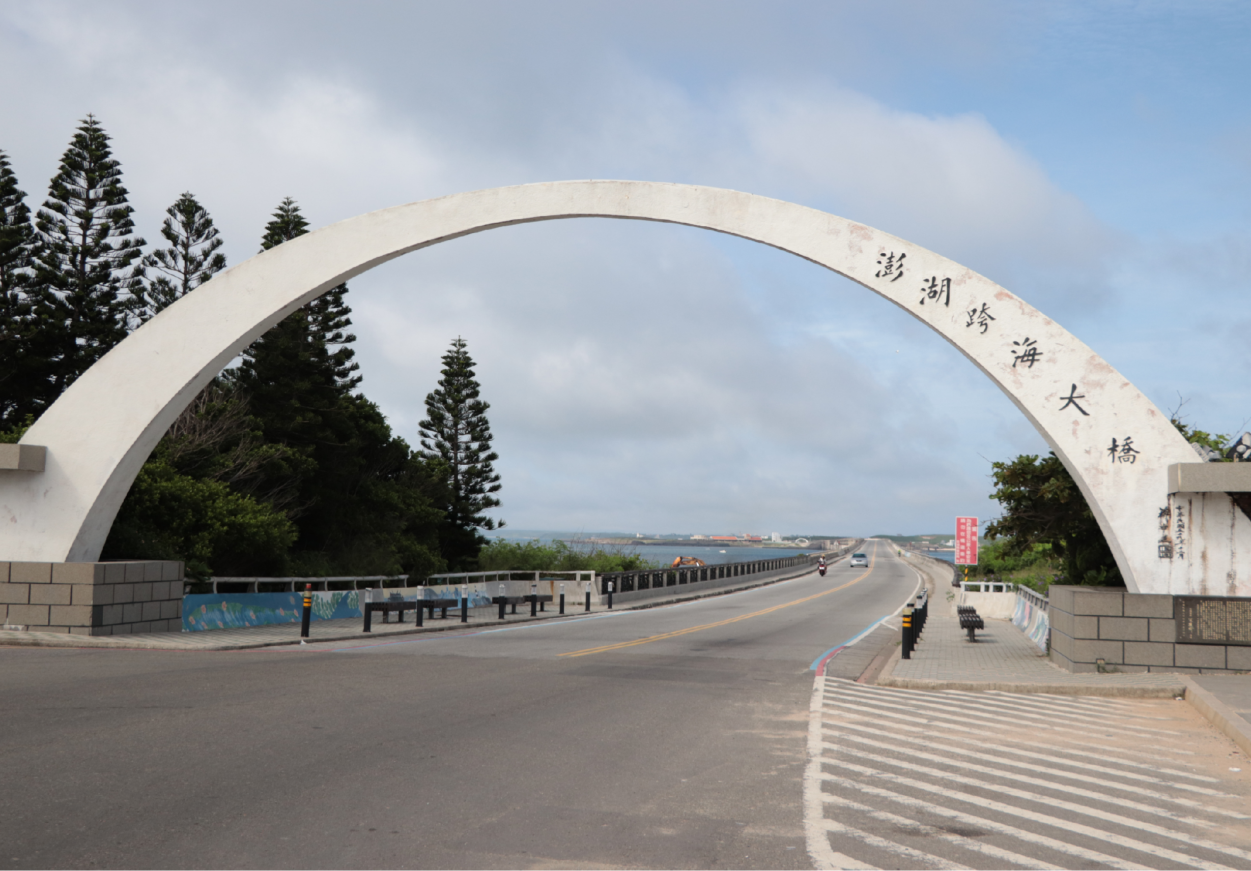 澎湖跨海大橋的白色拱形門牌，上刻有中文字。橋樑跨越海峽，連接澎湖本島與外島。橋下為寬闊的多車道公路，路旁種植多株蘇鐵樹。遠方可見海岸線和港口設施。天空晴朗，雲層稀疏。