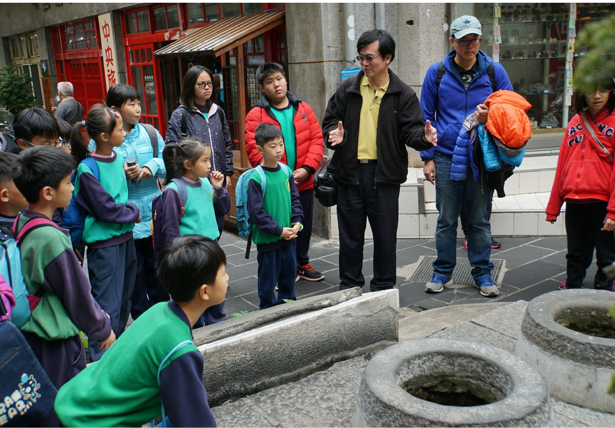 一群穿著綠色背心的小學生在街道上參加戶外教學活動，聚集在古井周圍聆聽成人講解員的介紹。背景是紅色傳統建築，展現地方文化教育的情景。