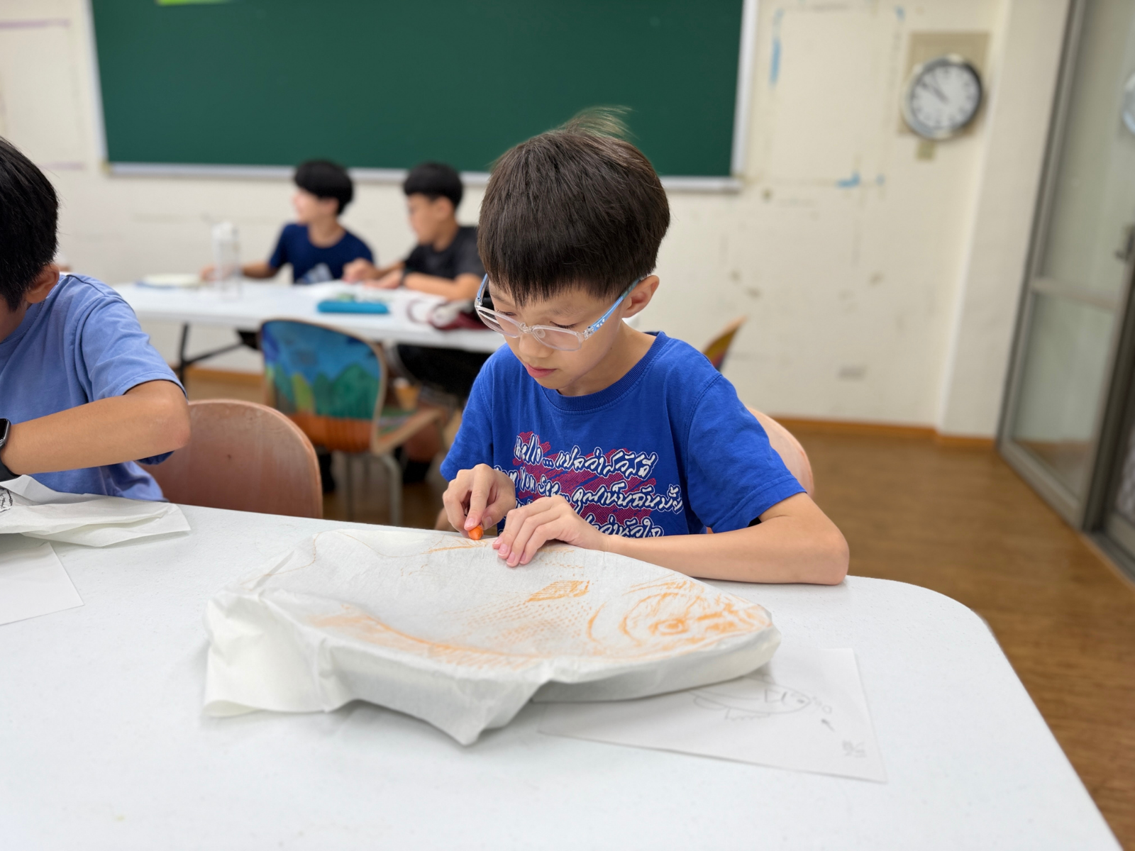 一位戴眼鏡、穿藍色上衣的男同學正坐在課桌前，手拿橘色蠟筆，專心致志地在覆蓋於魚體模型的宣紙上進行魚拓摩擦拓印。背景其他同學也在各自的座位上認真創作，教室裡瀰漫著靜謐的學習氛圍。