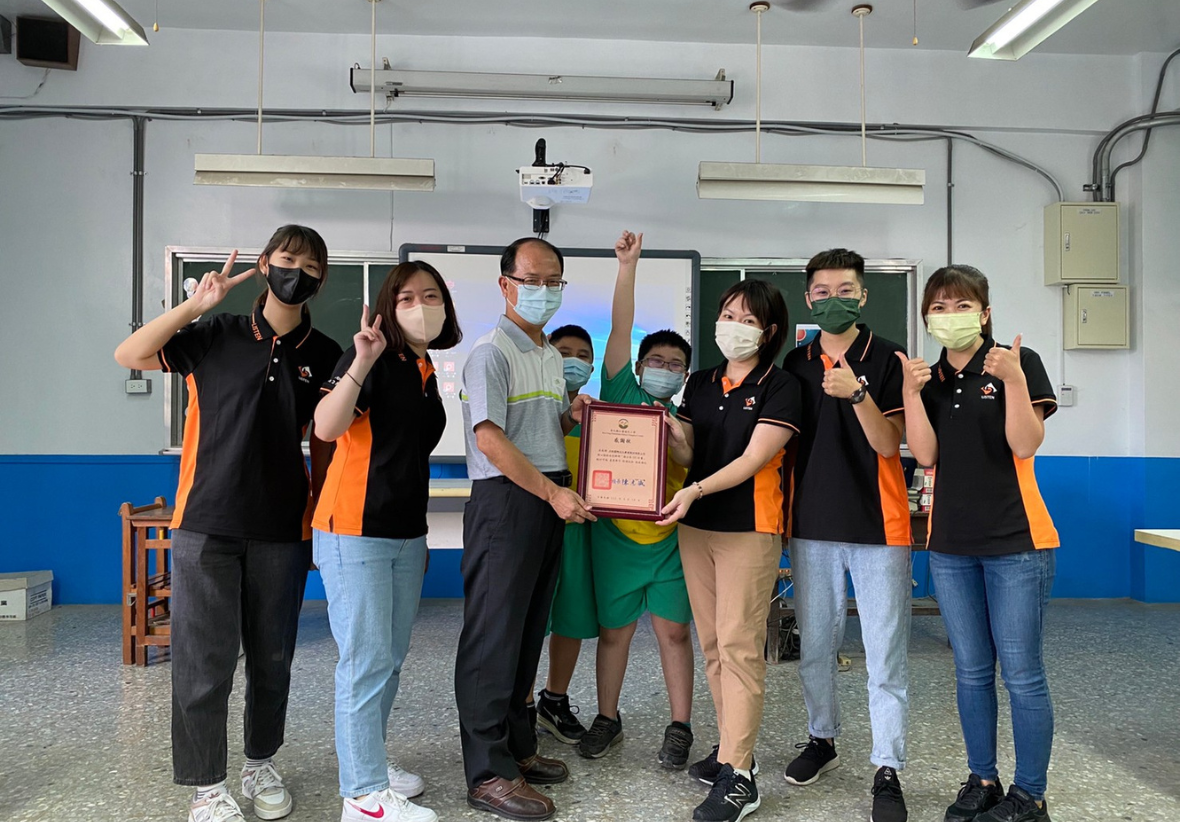 一張在教室內拍攝的課後團體合照。多位身穿黑橘雙色、印有公司標誌制服的講師，與學校師長及兩位戴口罩的小學生站在一起。師長與講師正共同手持一張裱框的感謝狀，大家開心地對鏡頭比著讚與勝利手勢。