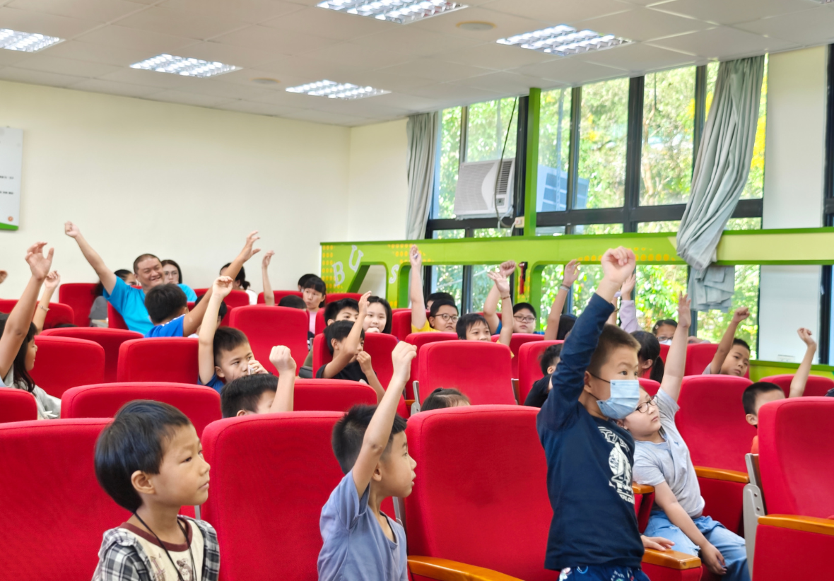 視聽教室內的課堂互動畫面。許多小學生坐在整齊的紅色沙發座椅上，每個人都非常熱烈、高高地舉起右手爭取回答問題的機會，台下展現出活潑且積極投入的學習氛圍。