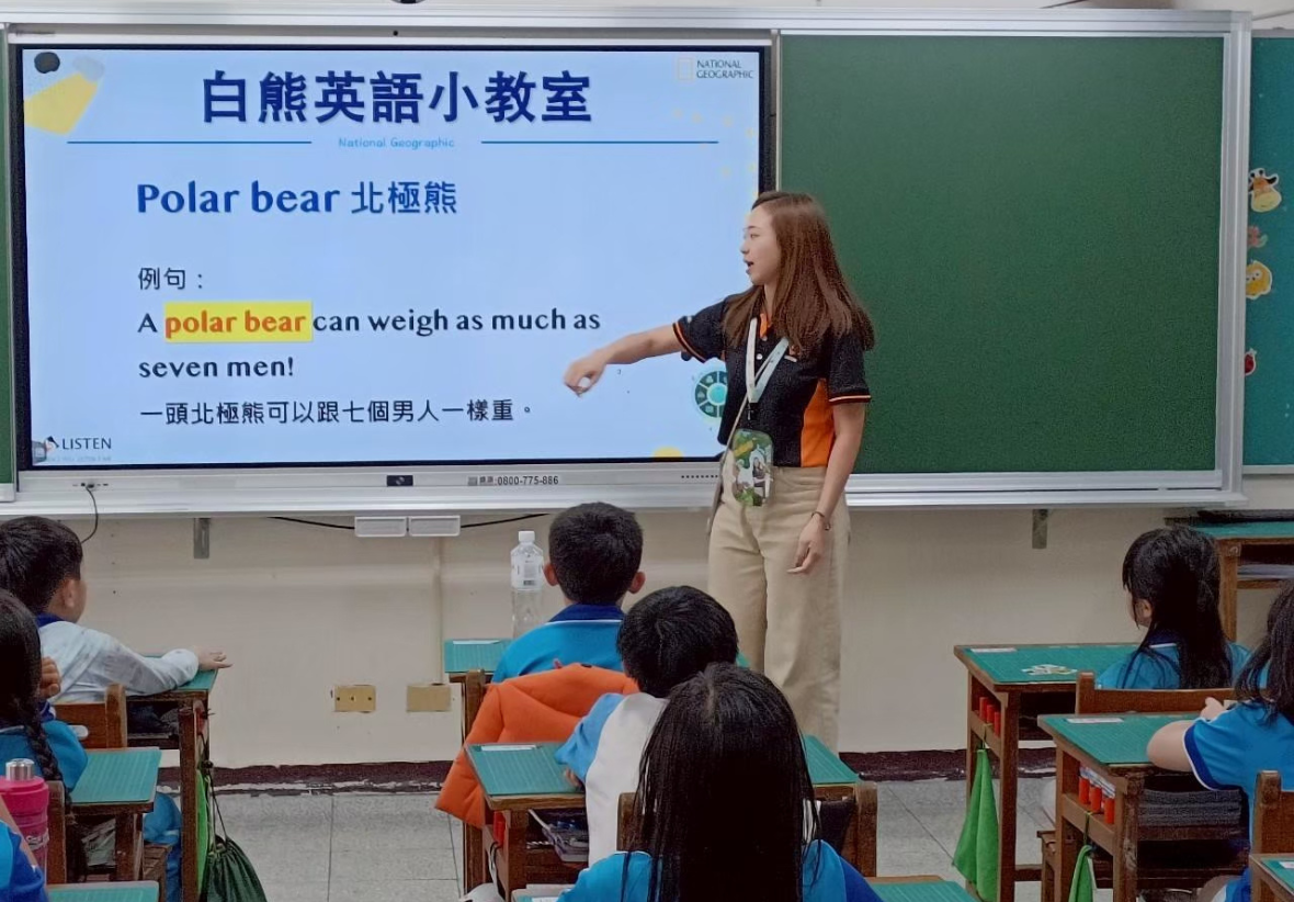 課堂教學現場。一位留著長髮、身穿黑橘制服的女性講師站在電子白板前，伸手指向螢幕上的「白熊英語小教室」簡報。簡報上寫著北極熊的英文單字與中文例句，台下的學生們則坐在課桌椅上專心聽講。