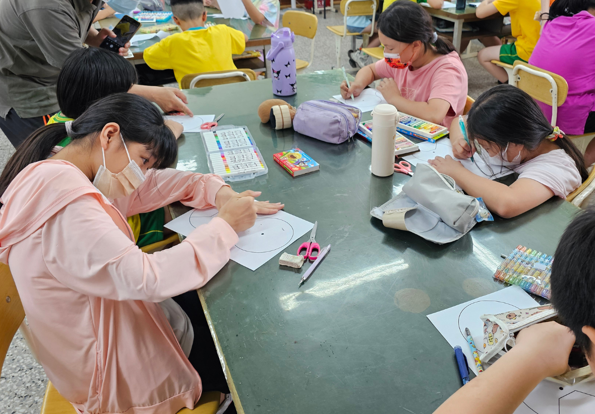 學生們進行手作教學的近距特寫。幾位戴著口罩的小學生圍坐在深綠色的課桌旁，桌上擺滿了彩色筆、蠟筆與剪刀。孩子們正專注地在印有圓形圖案的紙張上進行彩繪、裁剪與組裝。
