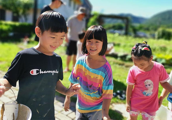 三個小孩在戶外農田場景開心笑著，左邊穿黑色Champion上衣的男孩，中間穿彩虹條紋衣的女孩，右邊穿粉紅色上衣的小女孩。背景是綠色草地和山景，其他大人在採收作物。