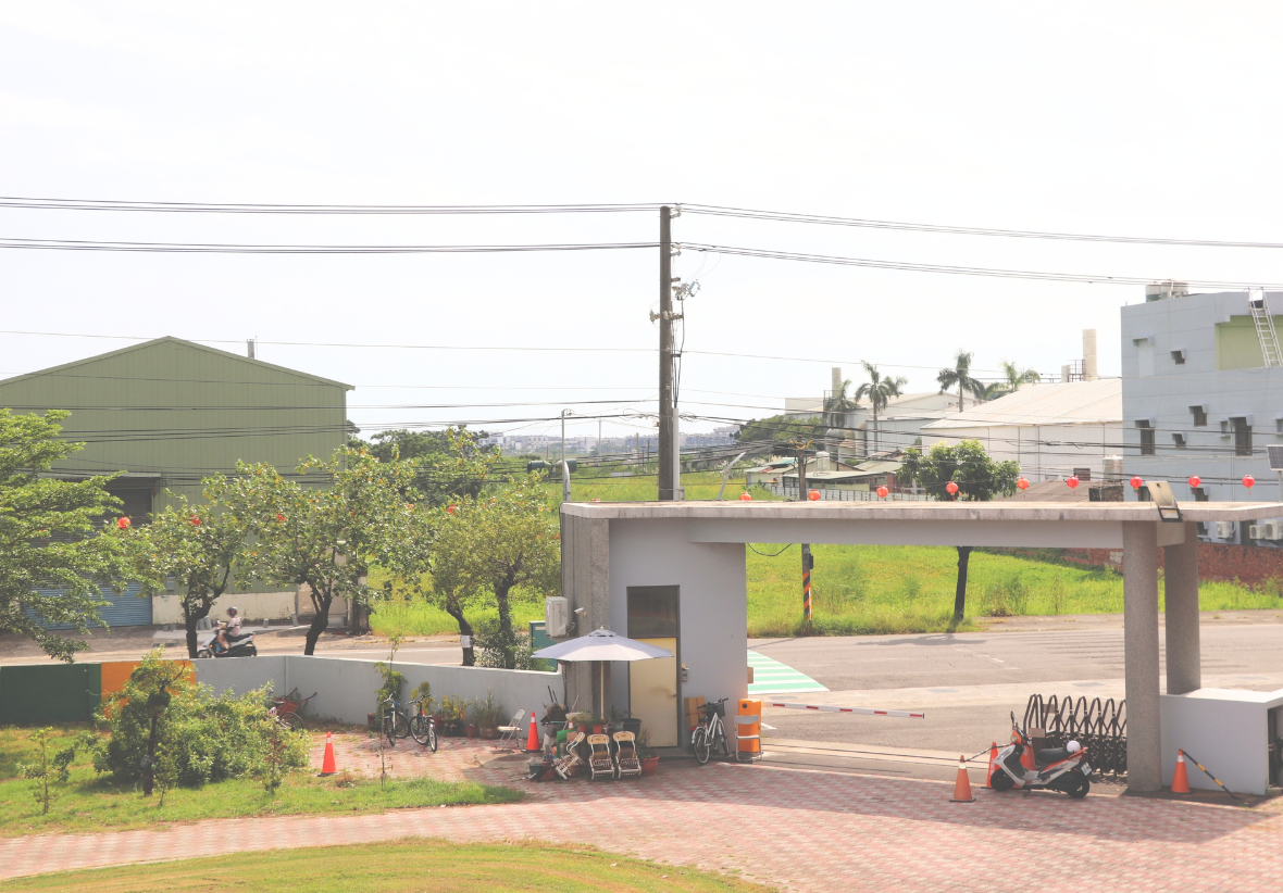 此圖為三侯國小的大門與警衛室視角，開闊的入口鋪設紅磚地面，設有電動柵欄與遮陽傘，門外為道路與遠處的平房、綠色田野景緻。