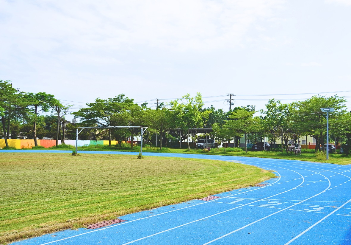 廣闊的校園操場與藍色PU跑道，跑道劃有1、2、3跑道線並延伸至遠方，中央為綠意盎然的草地足球場，周圍環繞著茂密的綠樹。