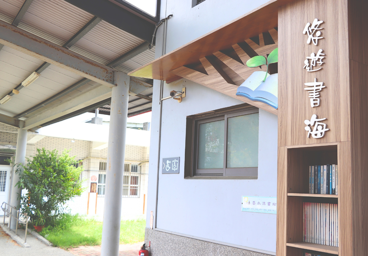 圖片為校園內的閱讀區一隅，木質柱體設置開放式書架與「悠遊書海」字樣及立體書本圖案，旁邊為穿堂與綠意盎然的庭園。