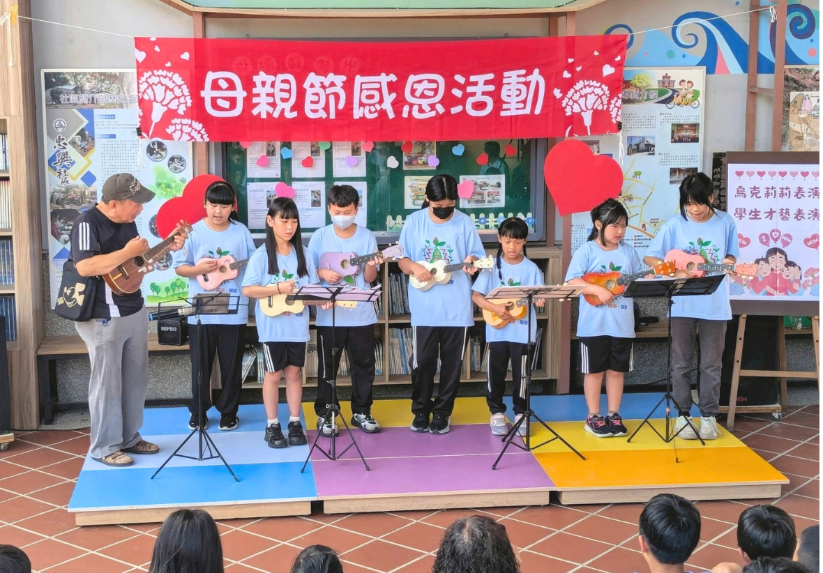 此圖為學校母親節感恩活動現場，背景掛有紅底白字布條，台上幾位小學生與老師手持烏克麗麗在舞台上合奏演出，台下有許多學生與家長認真觀賞。