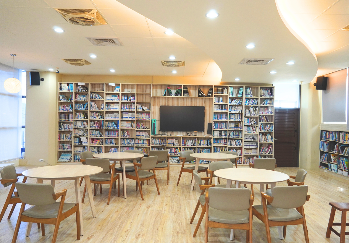 此圖為三侯國小的圖書館內景，天花板設計呈流線型，設有多組木質圓桌椅，後方整面牆為大型藏書櫃與電視螢幕，提供充實的閱讀空間。