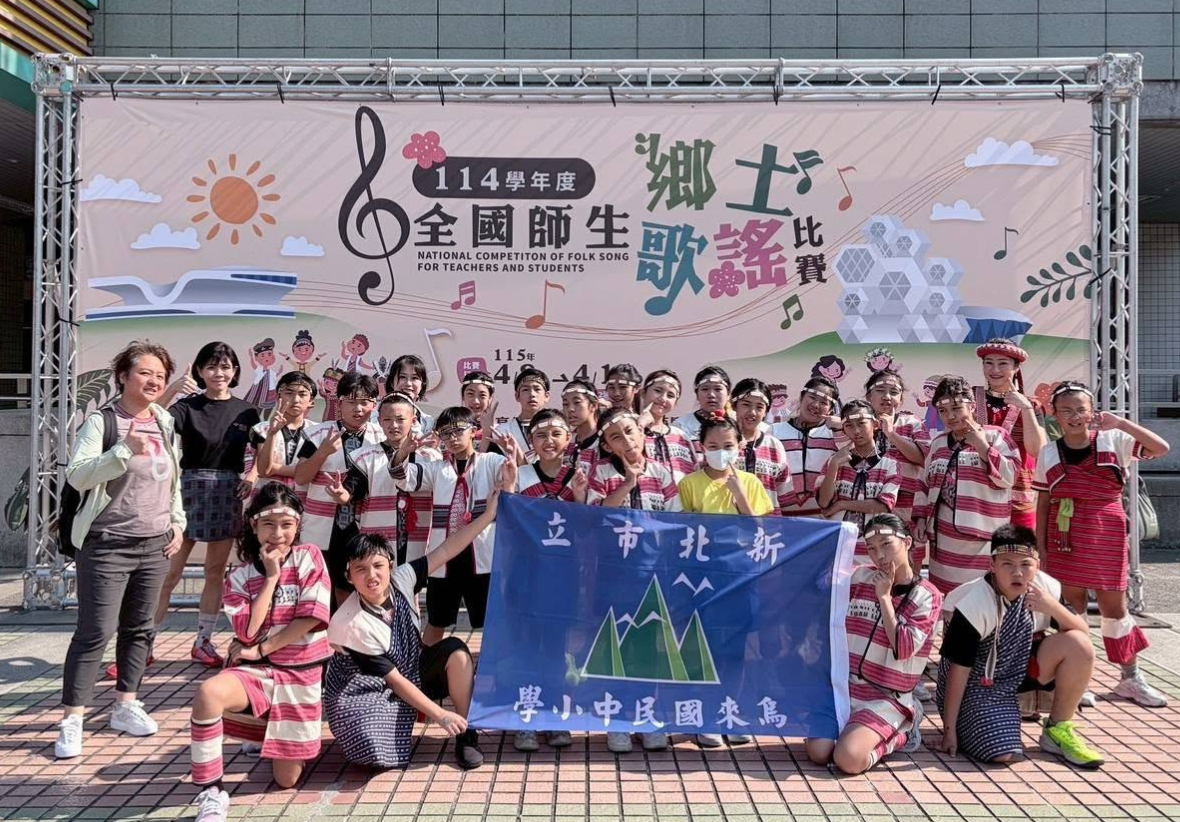 師生們在一面巨大的活動背板前開心大合照。背板上寫著「114學年度 全國師生鄉土歌謠比賽」。前排兩位同學拉著藍色的「新北市立烏來國民中小學」校旗，後方站滿了穿著泰雅族傳統服飾、面帶笑容並比出勝利手勢（V）的參賽隊員與指導老師。