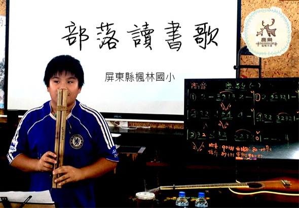 屏東縣楓林國小音樂教育活動現場，一位男性講師穿著藍色運動服，吹奏竹製樂器進行示範教學。背景投影幕展示「都巒讀書歌」標題，黑板上寫有樂譜和教學內容，講台上擺放傳統樂器。