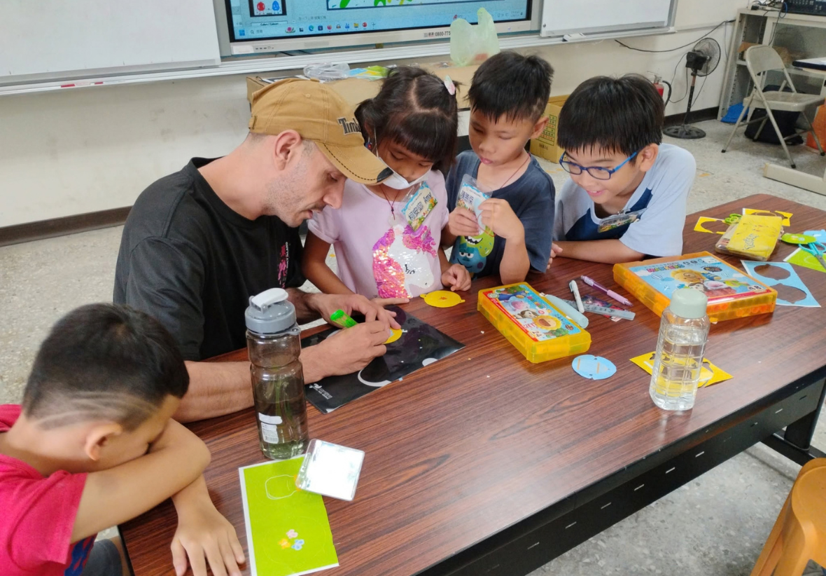 在教室課桌前，一位戴著鴨舌帽的外籍男志工正傾身指導三位國小學童進行美術手作。桌上擺滿了彩色筆、剪刀與勞作材料，孩子們圍在一旁認真觀察並學習，現場充滿溫馨的跨文化互動。