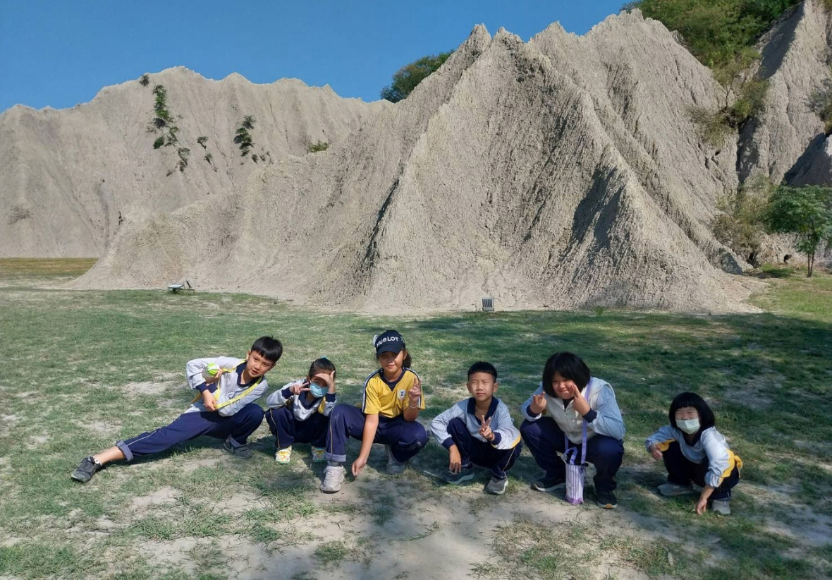 七位國小學童蹲坐在草地上，以田寮著名的泥岩惡地（月世界地貌）為背景開心合影。孩子們比著勝利手勢與俏皮動作，背後聳立著紋理清晰的灰白色泥岩山丘，突顯在地獨特的自然地質景觀。
