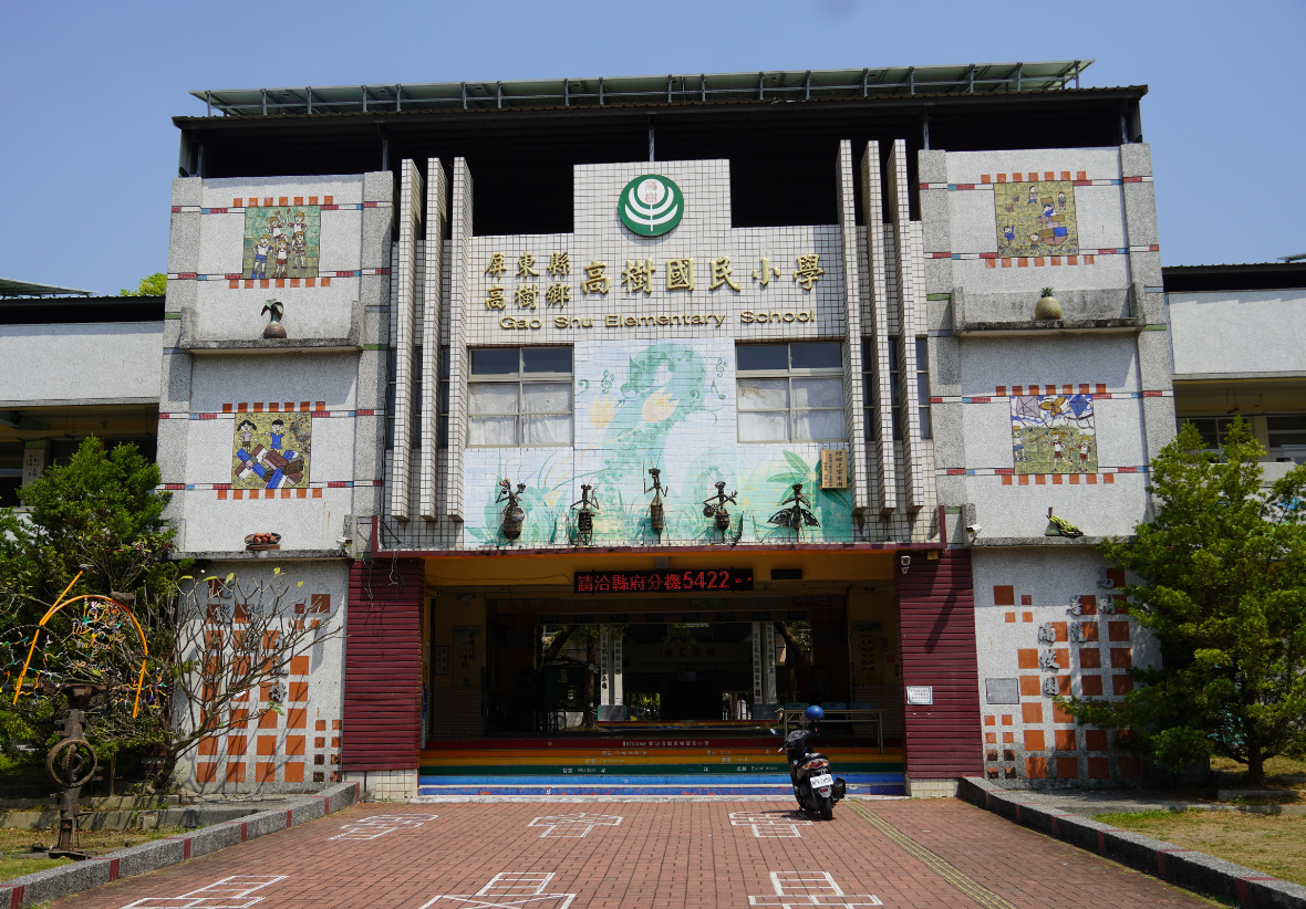 一所學校的正面建築，白色主體配紅色入口，牆面裝飾著橙色和彩色圖案。建築頂部有綠色校徽和中文校名。紅磚地面前方停放一台機車，兩側綠化樹木環繞，藍天晴朗。