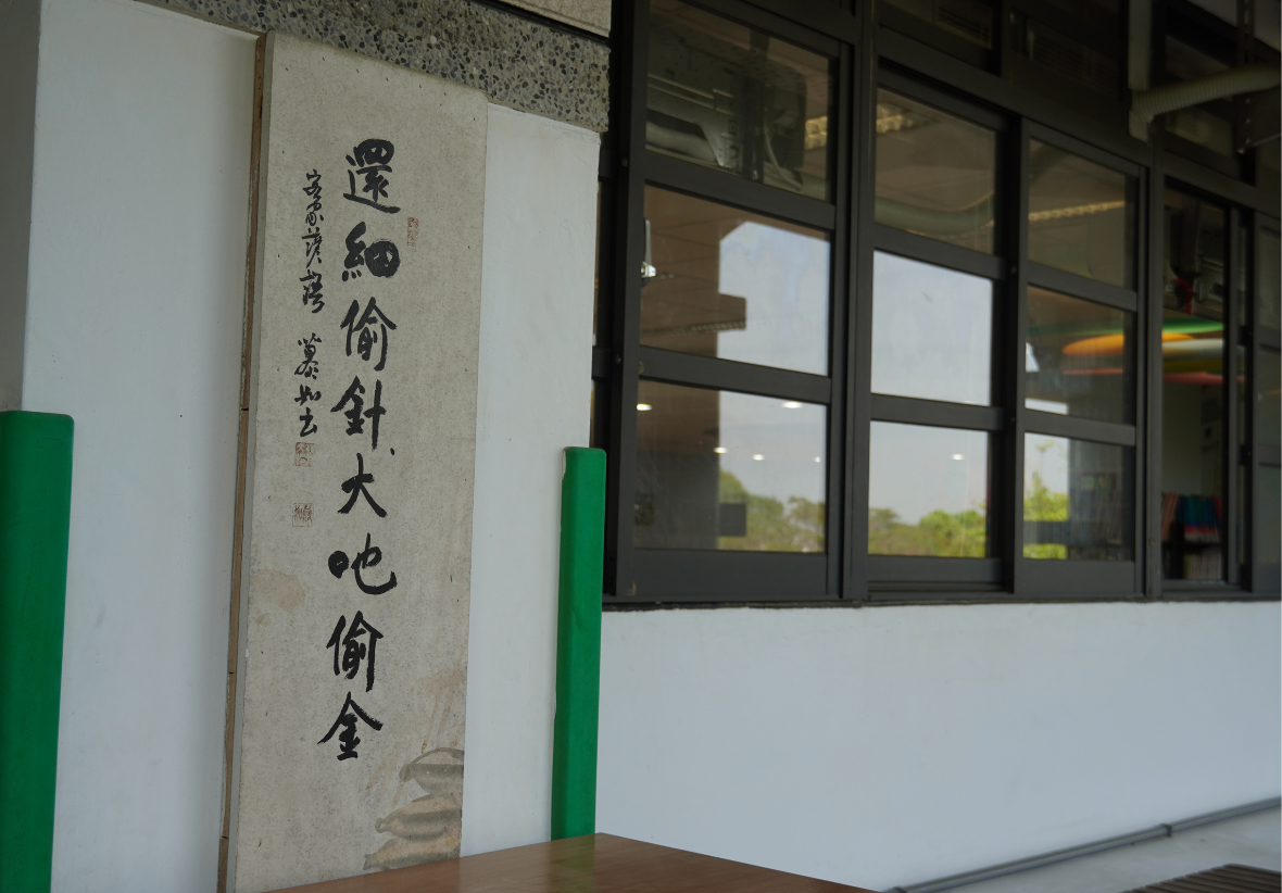 建築物入口處展示古文書法，寫有「還細偷針大也偷金」，為黃色底布上的黑色毛筆字，右方為玻璃門窗，體現傳統文化氛圍。