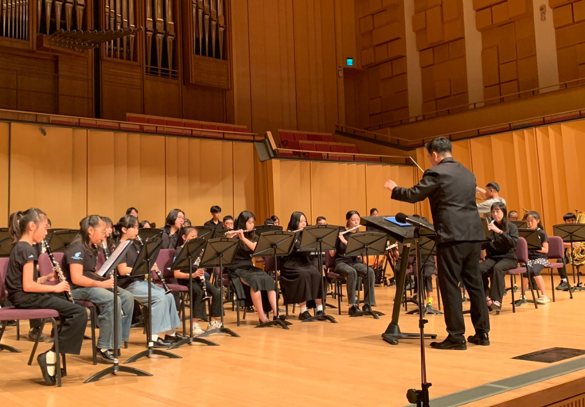 學生樂團在舞台上進行音樂演奏。約二十位穿著黑色制服的學生演奏各種樂器，由指揮者帶領。舞台背景有管風琴，牆面為木頭色。