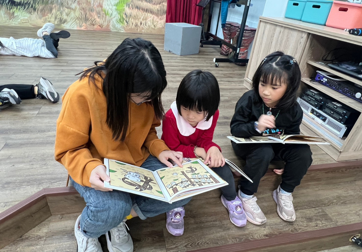 室內木質地板上，一位穿著橘色上衣的陪讀者正指著繪本內容，帶領兩位專注的小女孩一起閱讀。畫面左後方可以看到有其他孩子輕鬆地趴在地上看書，整體氣氛自由、舒適且充滿學習動力。