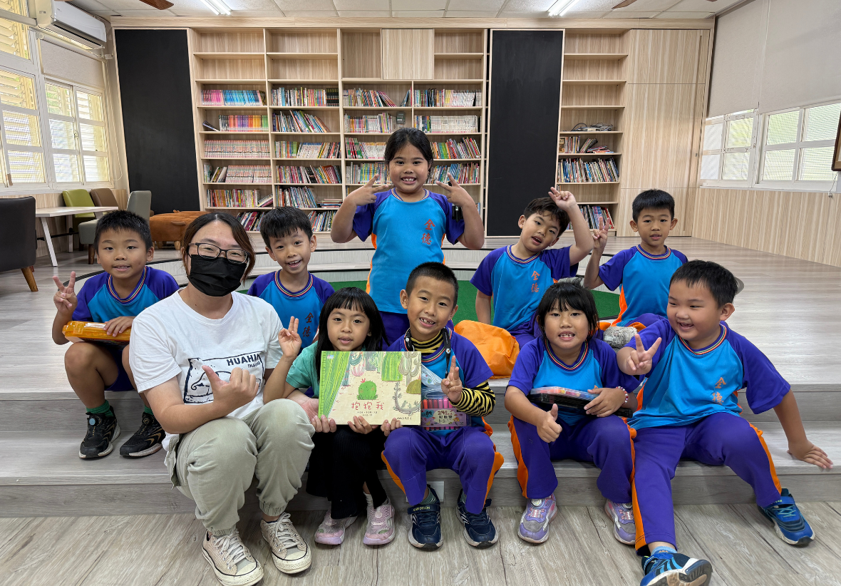 在一間大書櫃前，一位戴眼鏡的年輕女老師與十位穿著紫色與藍色運動服的小朋友開心合影。孩子們有的比出「V」手勢，有的舉起繪本《抱抱我》，每個人臉上都洋溢著燦爛的笑容，展現出偏鄉校園的朝氣。