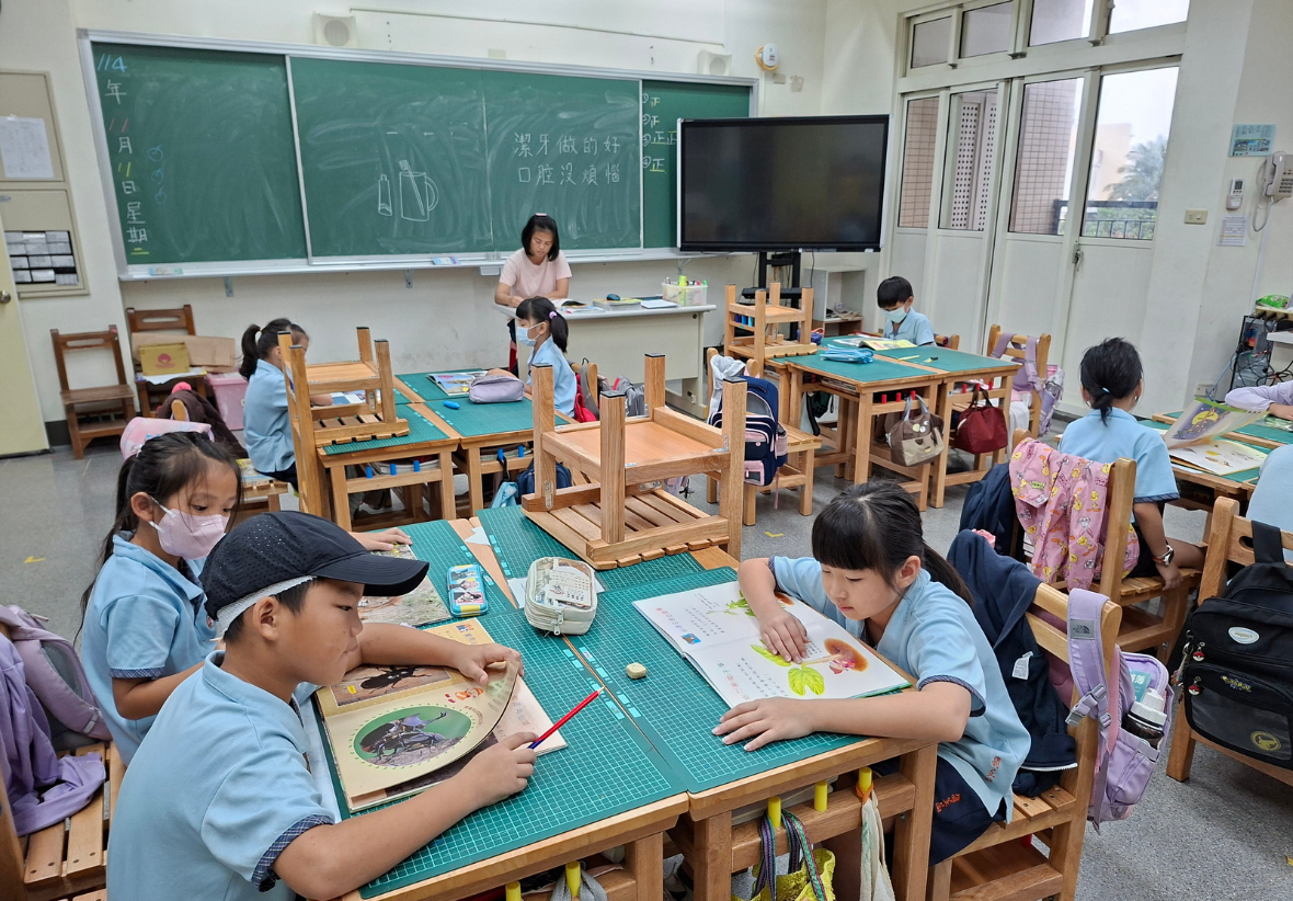 這是一張教室全景圖。穿著淺藍色制服的孩子們兩兩對坐，每個人桌上都放著精彩的繪本。有的孩子正在專心翻閱，有的正互相討論書中插圖。黑板上寫著「潔牙做得好，口腔沒煩惱」等生活叮嚀，呈現出日常校園學習的一面。