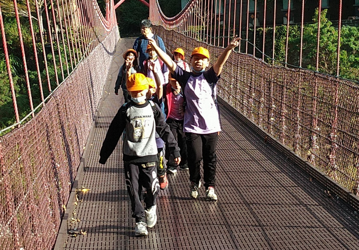 一群孩子正在通過吊橋，其中一位孩子高舉雙手比YA，看起來很開心。