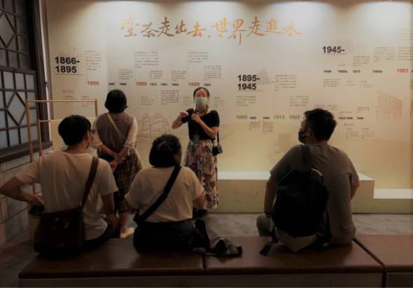 博物館內遊客參觀歷史展覽牆面，展示1866至1945年間的時間軸，分為1866-1895年和1895-1945年兩個時期，牆上有中文標題和詳細的歷史文字介紹，一名講解員向遊客說明展覽內容。