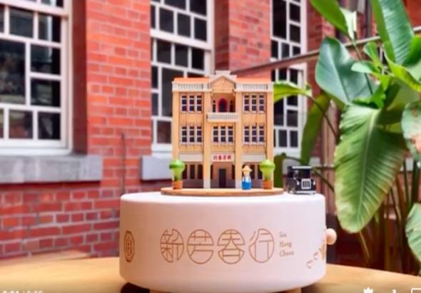 白色圓形旋轉台上展示一座黃色木製建築模型，為新芳春戲院的迷你版。背景為紅磚牆和綠色植物，展現懷舊建築與現代展示的對比。