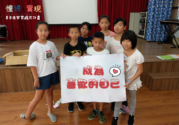 六名學童在室內禮堂舉行活動，手持寫有紅色中文字「成為愛我的自己」的白色海報牌，背景為紅色布幕。左上角有活動標誌，展示兒童關愛宣傳主題。